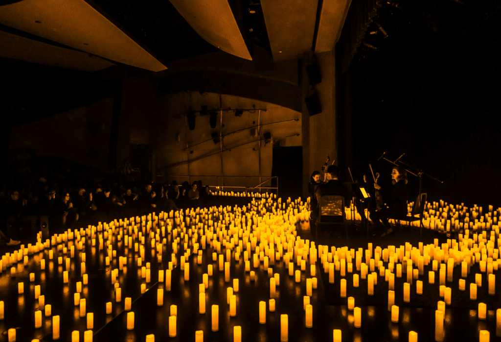 A photograph taking from the side of a stage showing sea of candles covering the stage with a string quartet performing for an audience.