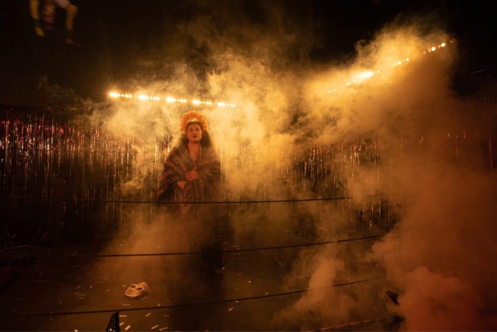 A masked woman stands in a smoky wrestling ring