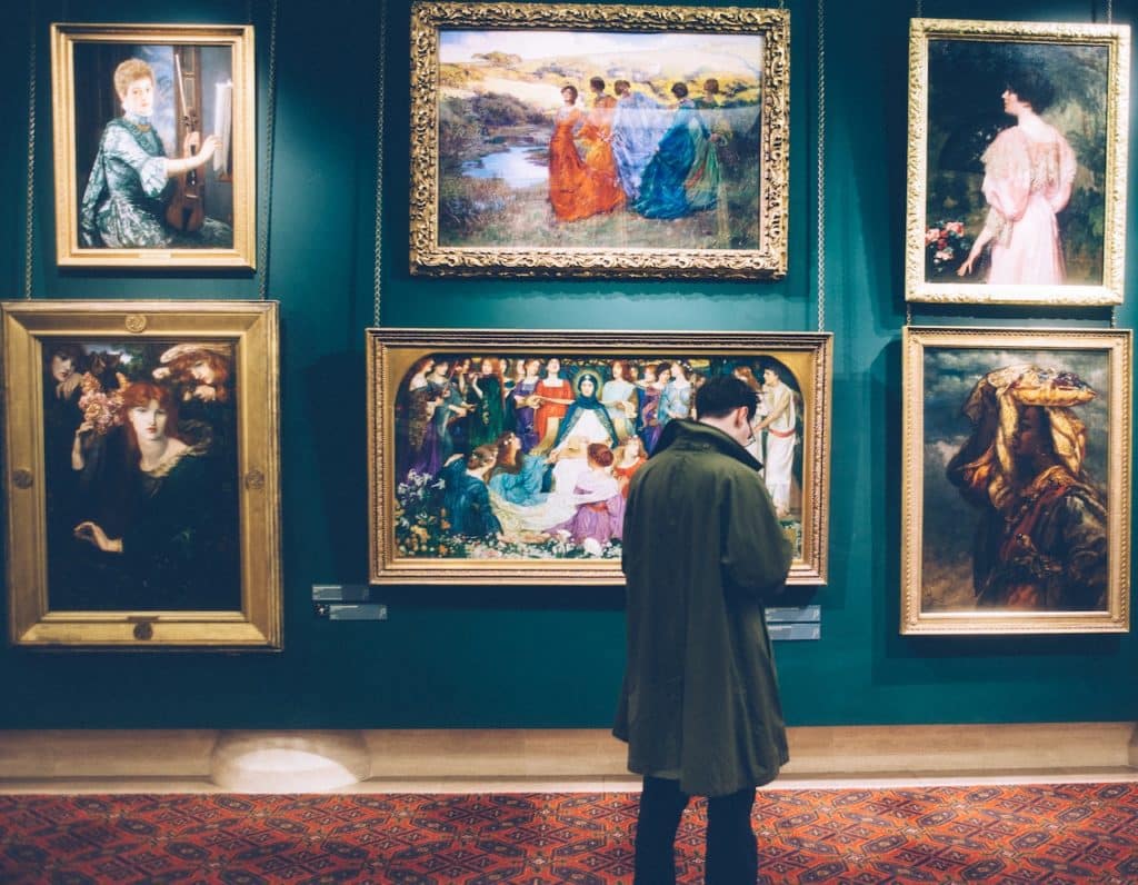 A man in a trench coat standing in front of several romantic paintings in a museum